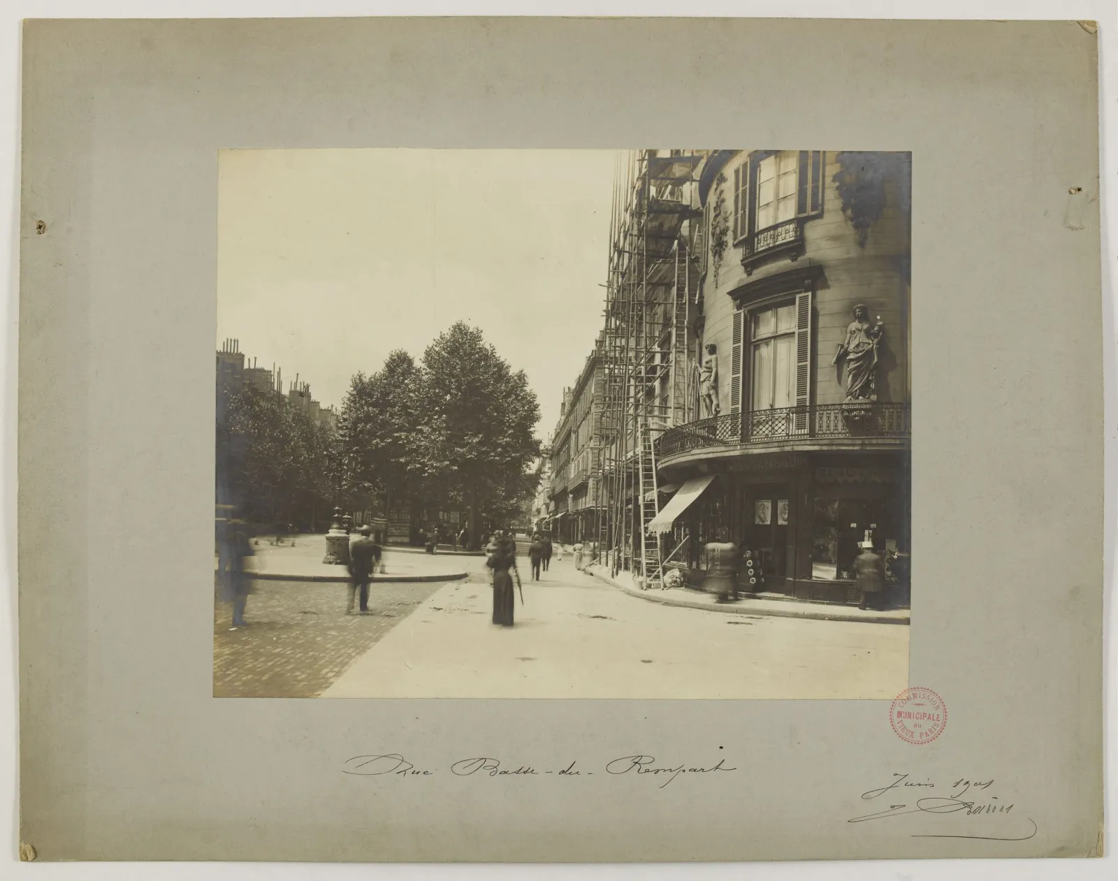 Mounted sepia photograph of a Paris street, with pedestrians, trees and scaffolding beside buildings; handwritten title, June 1901 date and a red archival stamp appear below.